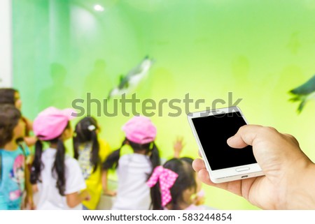 Man use mobile phone, blur image of children look at penguins in a zoo as background.
