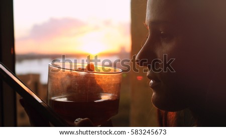 Young woman uses the phone, standing at the window drinks coffee on the background of a beautiful sunset with sun flare