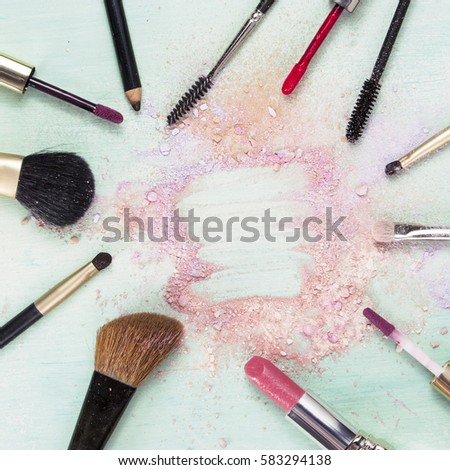 Makeup brushes and lipstick on teal blue background, with traces of powder and blush, forming a frame. Square template for makeup artist's business card or flyer design, with copyspace