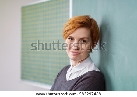 Portrait of young pretty woman with short haircut in dark sweater leaning back at green chalkboard in classroom, looking at camera