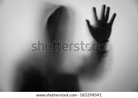 Shadow blur of horror man in jacket with hood.One hands on the glass.Dangerous man behind the frosted glass.Mystery man.Halloween background.Black and white picture.Blur picture