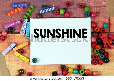 A happiness concept image of a colorful beads and cute clothespin scattered over a colorful wooden texture background with a white canvas and a word SUNSHINE