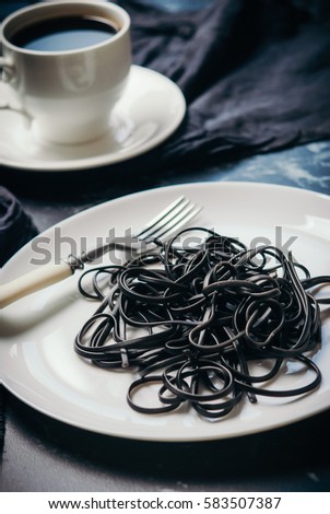 black spaghetti with squid ink on a white plate and a cup of hot black coffee on a black marble background. Concept black and white breakfast. Toned picture