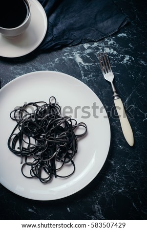 black spaghetti with squid ink on a white plate and a cup of hot black coffee on a black marble background. Concept black and white breakfast. Toned picture. Top view