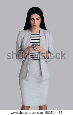 Using modern technologies. Beautiful young woman in smart casual wear using her smart phone while standing against grey background