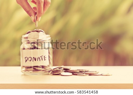 Hand putting Coins in glass jar with DONATE word written text label for giving and donation concept Royalty-Free Stock Photo #583541458