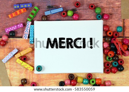 A happiness concept image of a colorful beads and cute clothespin scattered over a colorful wooden texture background with a white canvas and a word MERCI in french mean THANK YOU