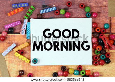 A happiness concept image of a colorful beads and cute clothespin scattered over a colorful wooden texture background with a white canvas and a word GOOD MORNING