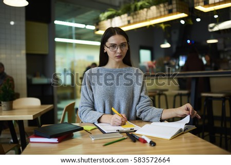 Concentrated female skilled excellent student working on coursework searching information for research analyzing scientific articles and textbooks learning to become best in modern cafe interior 
