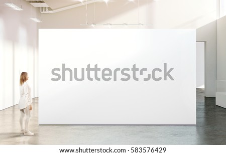 Woman walking near blank white wall mockup in modern gallery. Girl admires a clear big stand mock up in museum with contemporary art exhibitions. Large hall interior, banner exposition Royalty-Free Stock Photo #583576429