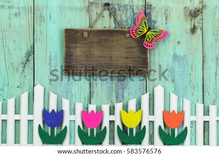 Blank wood sign with colorful butterfly hanging over white fence with purple, pink, yellow and orange spring flowers with rustic antique mint green background; springtime background with copy space