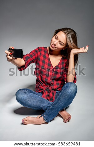Lovely girl wearing jeans and red checked shirt sitting barefooted at floor making self portrait on her smart phone, studio shot