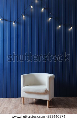 White fabric armchair on a background of blue wall with retro garland of light bulbs. Modern interior. Texture for the design