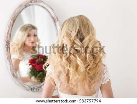 Beautiful curly haired bride in a wedding dress at a mirror in white room. Girl repeats the hairstyle and makeup