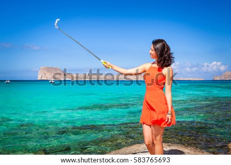 Pretty young model dressed in a coral clothes having fun making selfie with a selfiestick on the background of the clear blue sea