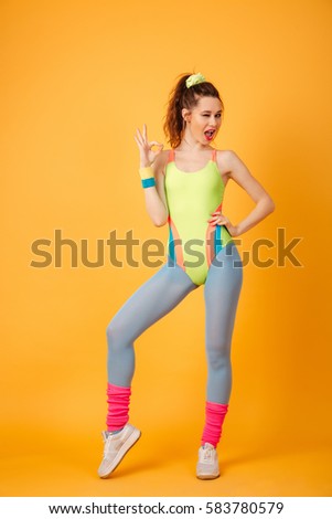 Picture of smiling young fitness lady posing over yellow background. Looking at camera make okay gesture.