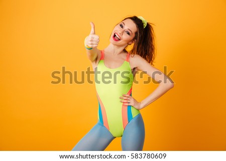 Picture of smiling young fitness woman posing over yellow background. Looking at camera make thumbs up gesture.
