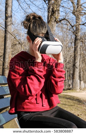 Young girl with glasses of virtual reality looks aside outside sitting on a bench