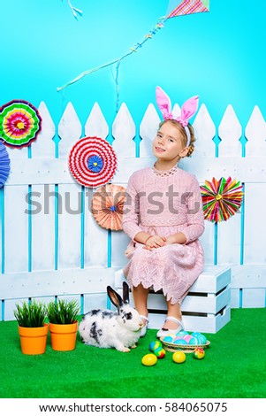Cute happy little girl sitting with Easter Bunny in spring decorations. Easter Bunny and painted eggs. Kid's fashion. 