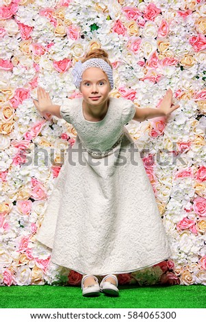Cute smiling girl in beautiful white dress standing by a floral background. Spring and summer inspiration. Kid's fashion. Full length portrait.