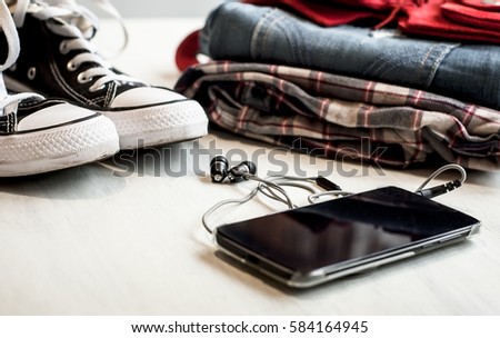 Travel accessories: hipster sneakers, denim shorts, plaid shirt, cowboy belt and phone with headphones on wooden background. Selective focus. Traveling concept