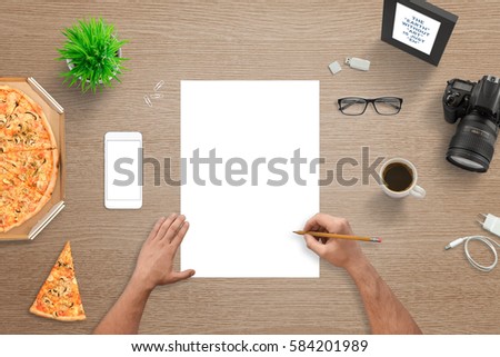 Man sketching on empty white paper. Top view of desk. White smart phone with blank screen for mockup. Digital camera, pizza, plant, glasses, coffee, photo frame beside.