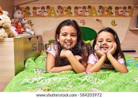 Two funny sisters lying on the bed and smiling in color nursery.