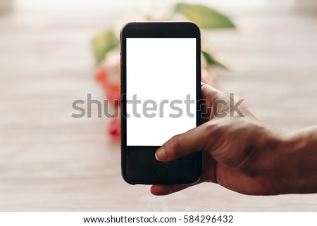 hand holding phone with empty screen with space for text above pink tulips on white wooden rustic background. instagram photographer, blogging workshop concept. 