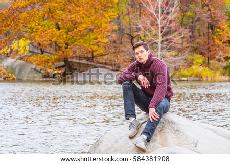 Young American man wearing purple shawl collar pullover knit sweater, black jeans, gray casual shoes, sits on rocks by lake at Central Park, New York in autumn day, relaxing. Color filtered effect