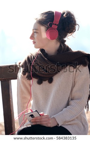 Smiling travel teenage girl with scarf and pink headphone sitting outside looking at mobile phone