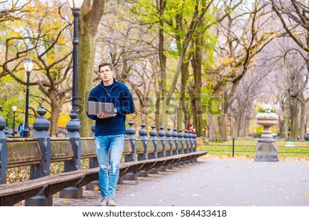 Way to Success. Man wearing blue sweatshirt, fashionable jeans, walks on road with long bench, trees at Central Park, New York in autumn day, reads, works on laptop computer. Color filtered effect
