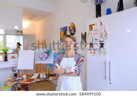 Cute female painter posing, laughing and fooling around in front of camera and smiling with brushes and colorful palette in hand, standing in studio with white art paintings on walls. Girl European