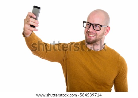 Studio shot of young happy bald muscular man smiling while taking selfie picture with mobile phone