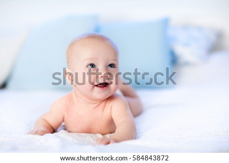 Baby boy wearing diaper in white sunny bedroom. Newborn child relaxing in bed. Nursery for children. Textile and bedding for kids. Family morning at home. New born kid during tummy time with toy bear Royalty-Free Stock Photo #584843872