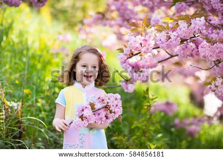 Little happy girl playing under blooming cherry tree with pink flowers. Child holding sakura blossom. Summer fun for family with kids outdoors in a beautiful spring garden. Kid with flower on Easter