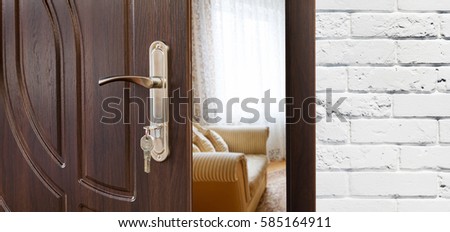 Half opened door handle closeup, entrance to a living room, sofa and window. Welcome, privacy concept. Door lock with keys, white brick wall, modern interior design.