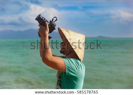 Young caucasian man in vietnamese traditional hat taking photo on a luxury tropical beach with turquoise water, blue sky. Samui island, Thailand.