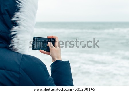 Close-up of a woman in a winter jacket with a hood using a mobile phone on the beach