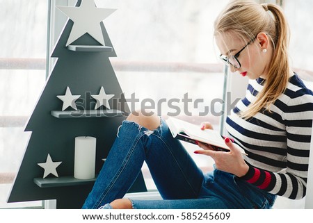 Blonde lady in black glasses reads book sitting before wooden Christmas tree on balcony