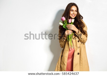 Fashion photo of a beautiful young woman with tulips in her hand .she dressed  in a beautiful brown trench coat, crimson dress.Spring concept. March 8. beautiful girl in stylish clothes.
