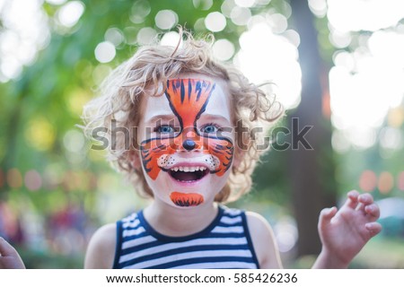 smiling boy with curly hair and face art painting like tiger, little boy making face painting, halloween party, child with funny face painting, little cute boy with faceart on birthday party close up Royalty-Free Stock Photo #585426236