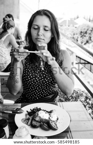Young woman taking picture of her breakfast made of poached eggs benedict served on toast with smoked salmon, avocado, grilled tomato and spinach, with smart phone 