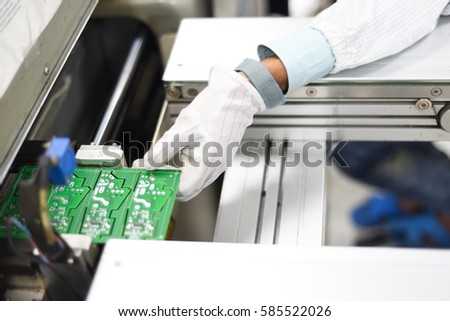 Man working in an electronics factory removing printing circuit board from SMT line. Image taken at Electronics manufacturing plant and the PCB board assembled with electronic components. Royalty-Free Stock Photo #585522026