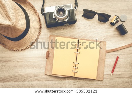 Man hand writing a notebook with film camera, wicker hat and sunglasses on wooden table - top view.