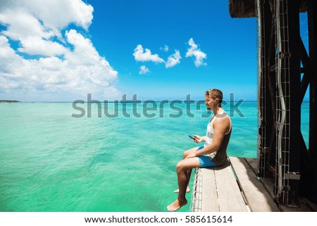 Young confident man sitting on pier taking pictures of wonderful tropical view, enjoying sunny day
