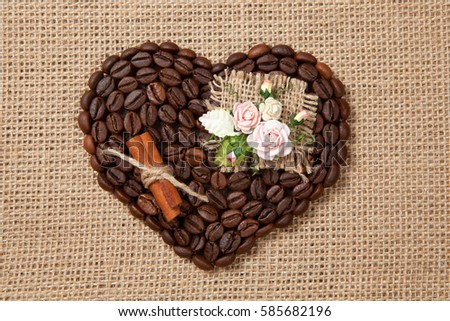Heart of coffee beans with artificial flowers and cinnamon stick on sackcloth.