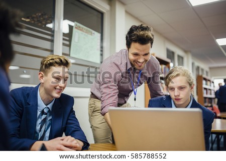 Teen students are working on a laptop in their school lesson. A teacher is leaning over them, helping them with some work.  Royalty-Free Stock Photo #585788552