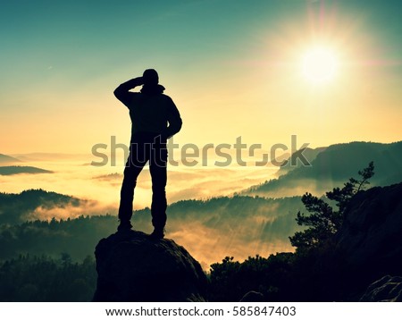 Man silhouette climbing high on cliff. Hiker climbed up to peak enjoy view. Man  watch over misty and foggy morning valley in morning Sun.  Royalty-Free Stock Photo #585847403