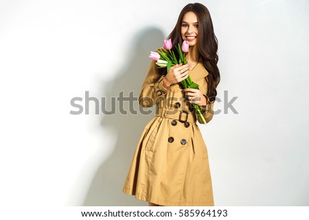 Fashion photo of a beautiful young woman with tulips in her hand .she dressed  in a beautiful brown trench coat, crimson dress.Spring concept. March 8. beautiful girl in stylish clothes.


