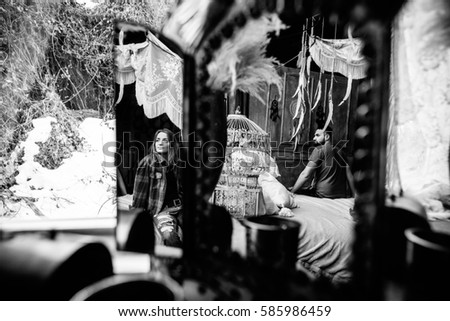 Reflection of the woman in the mirror, she looks out the window. Man sitting on a bed and looking at his woman. Black and white photo, reflection. Love story